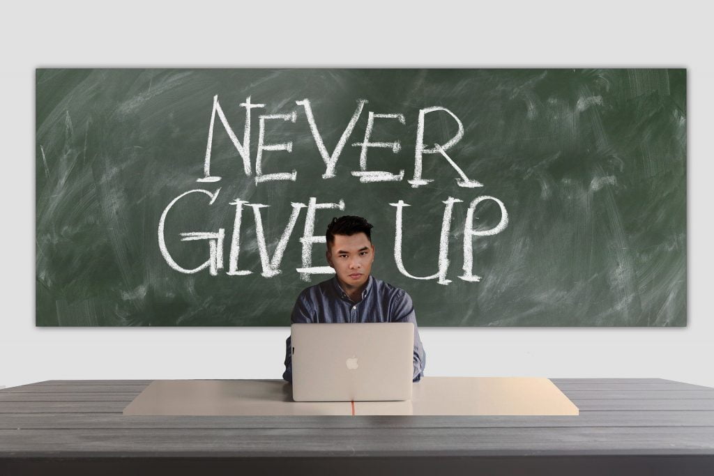 An Asian man sitting behind a desk with a laptop with a blackboard behind him with "Never Give Up" written on it